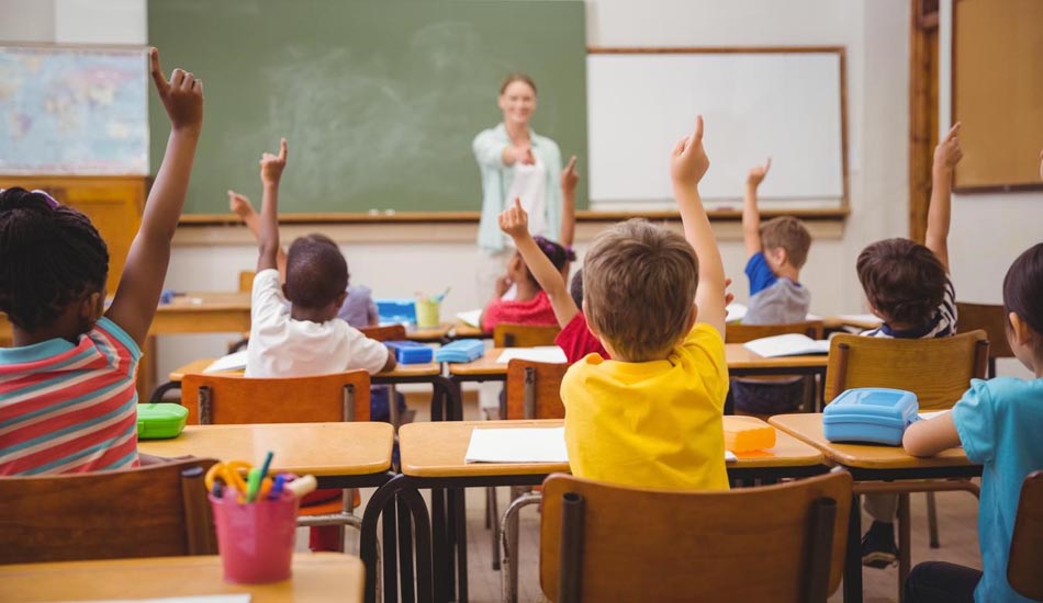 A female teacher points to one of her students who are all raising their hands in a classroom