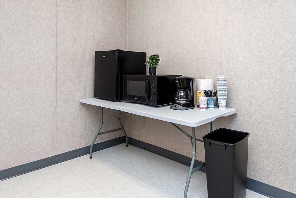 Breakroom table with mini fridge, microwave, coffee machine, coffee supplies, and trash can.