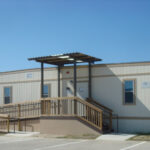 A beige modular building with a wooden ramp and stairs leading to the entrance.