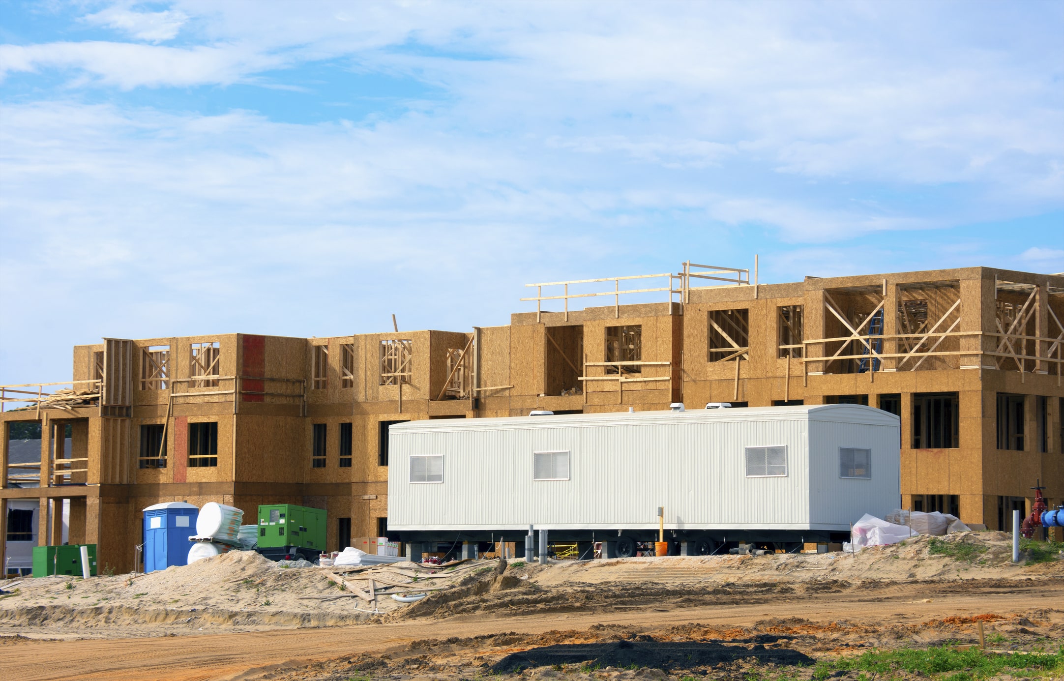 Large construction site half constructed wooden building with mobile job location office trailer in front area for contractors, work managers and supervisors for paperwork and worker interviews.